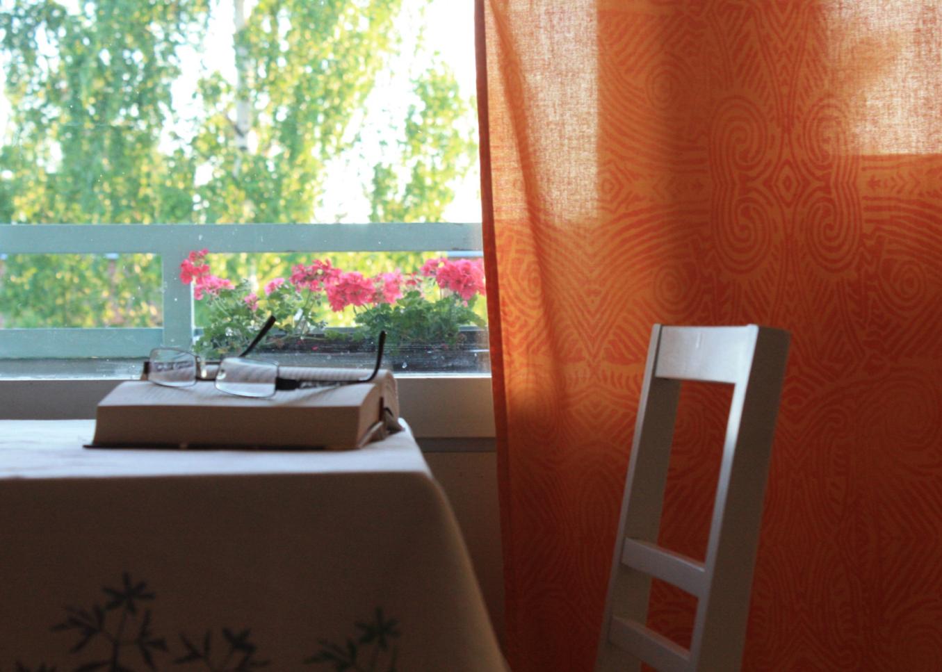 Vaalealla liinalla peitetty pöytä, jolla vanha kirja ja sen päällä silmälasit. Taustalla ikkuna josta näkyy vihreä kesämaisema. Ikkunassa kirkkaan oranssit verhot.